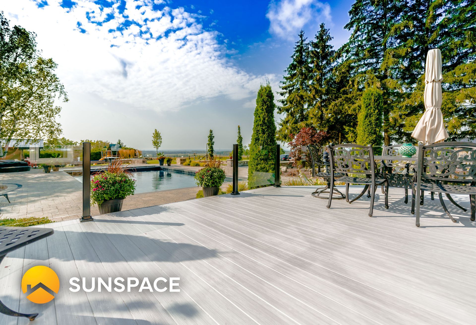 White composite deck with outdoor furniture, overlooking a pool and trees. Blue sky.