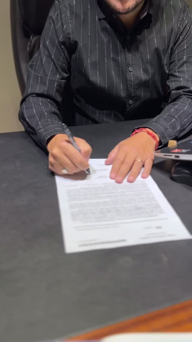 Persona firmando un documento en un escritorio, vestida con una camisa negra.