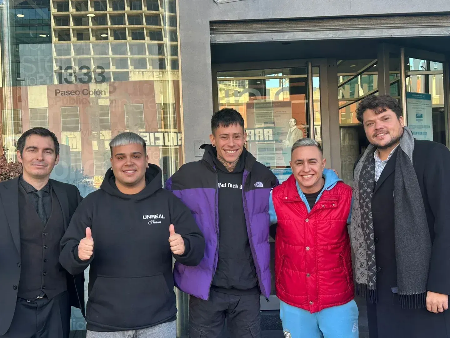 Cinco personas fuera de un edificio; una levanta el pulgar, la persona del medio sonríe y dos están abrazadas.