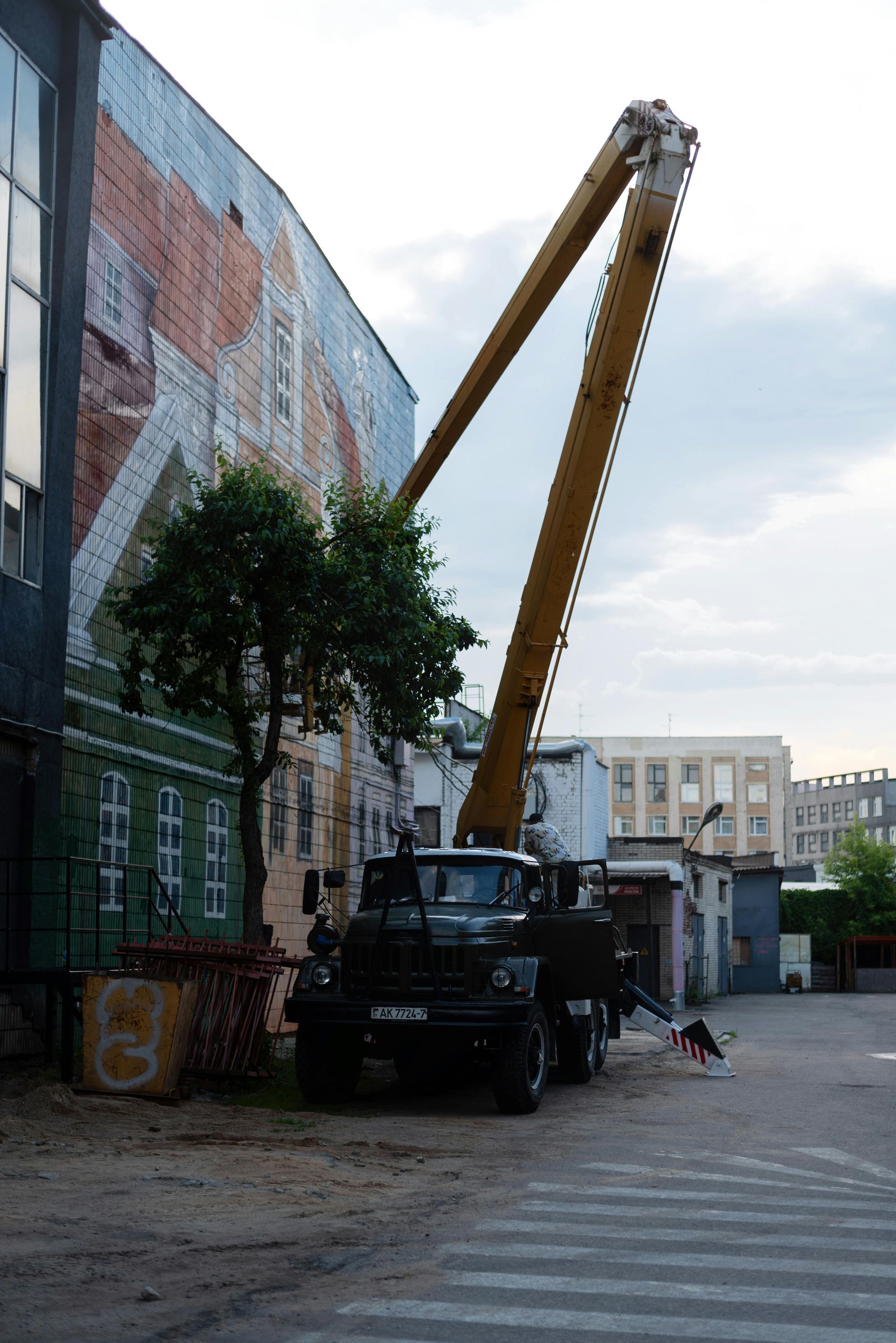 Truck with a tall boom extended toward a building with a mural. A small dog walks nearby.