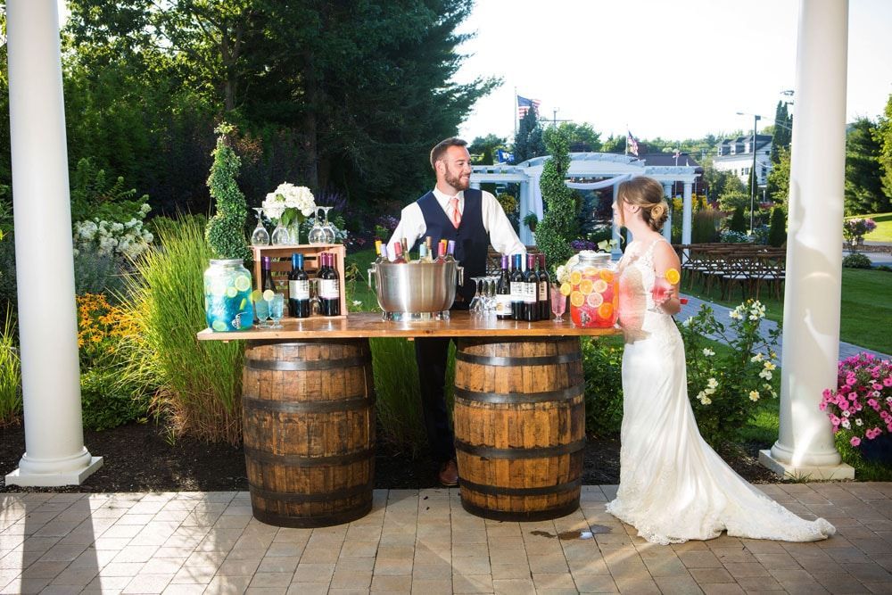 Bartender serves a bride at a rustic outdoor bar made of barrels. They're in a garden setting with white columns.
