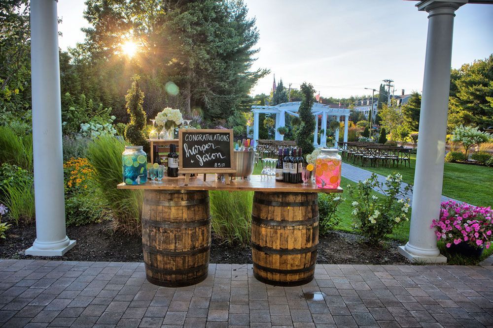 Outdoor bar made of barrels, with drinks, flowers, and a chalkboard sign, set against a garden with a sunny backdrop.