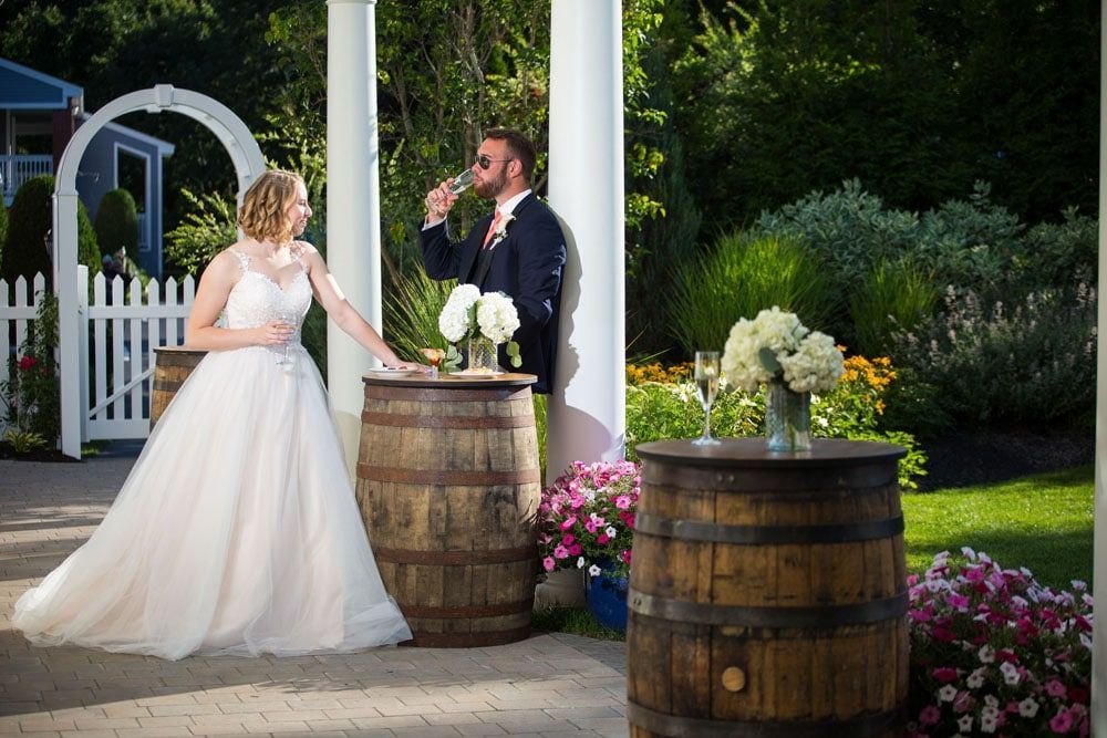 Bride and groom at an outdoor wedding reception. Bride stands by a barrel, groom sips champagne.