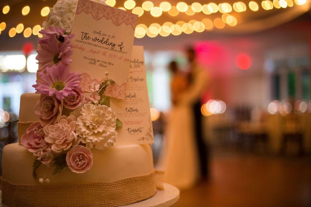 Wedding cake with flowers, burlap, and a menu card in front of a blurred couple embracing under string lights.