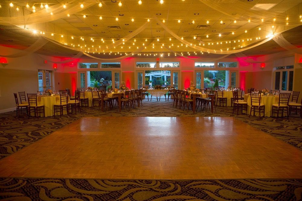 Wedding reception hall with dance floor, tables set with yellow cloths, string lights.
