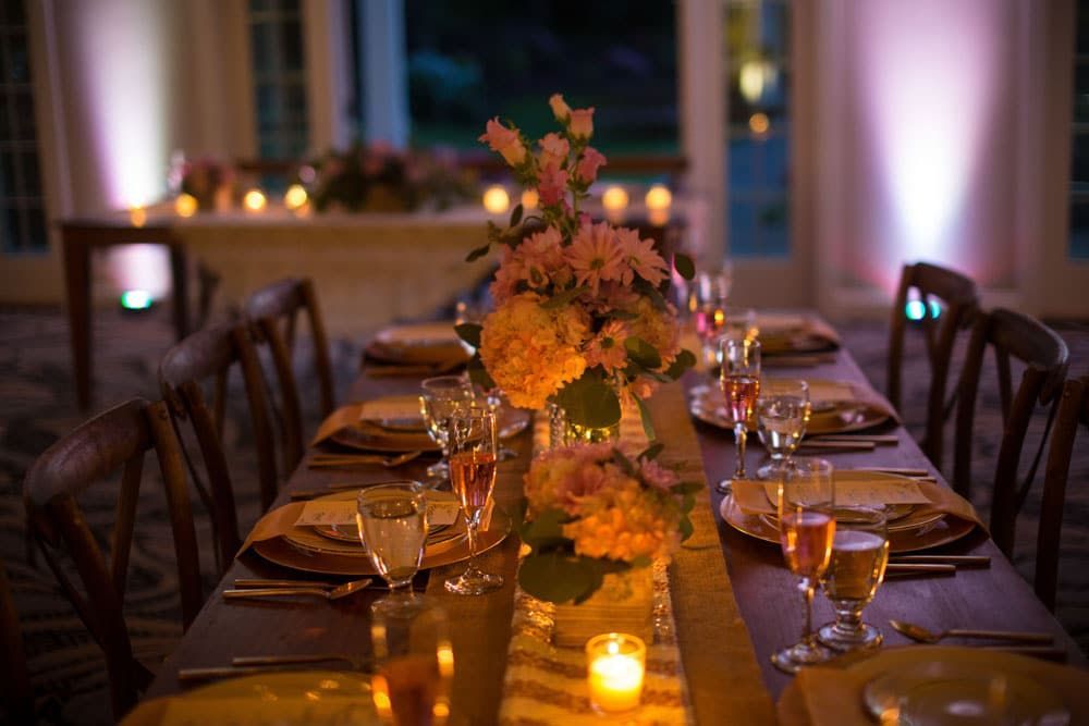 Long wooden table set for a formal dinner, with floral centerpiece and lit candles; interior setting.