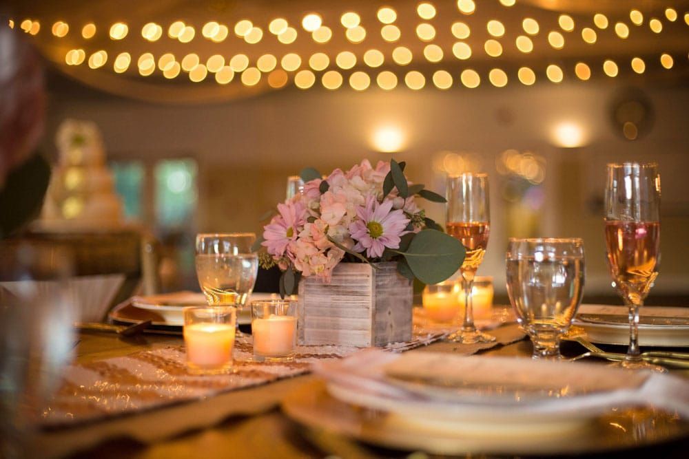 Wedding reception table with flowers, candles, and champagne glasses, with string lights in the background.