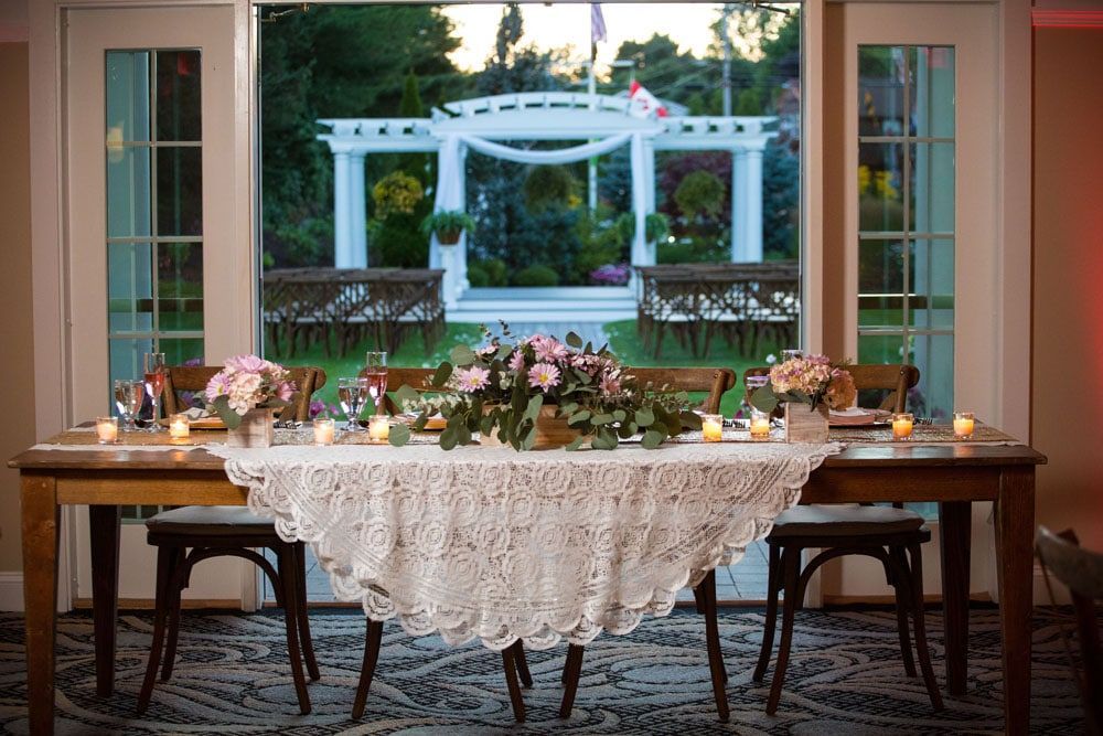 Wedding reception table set in front of French doors overlooking a garden with an arbor; flowers and candles.
