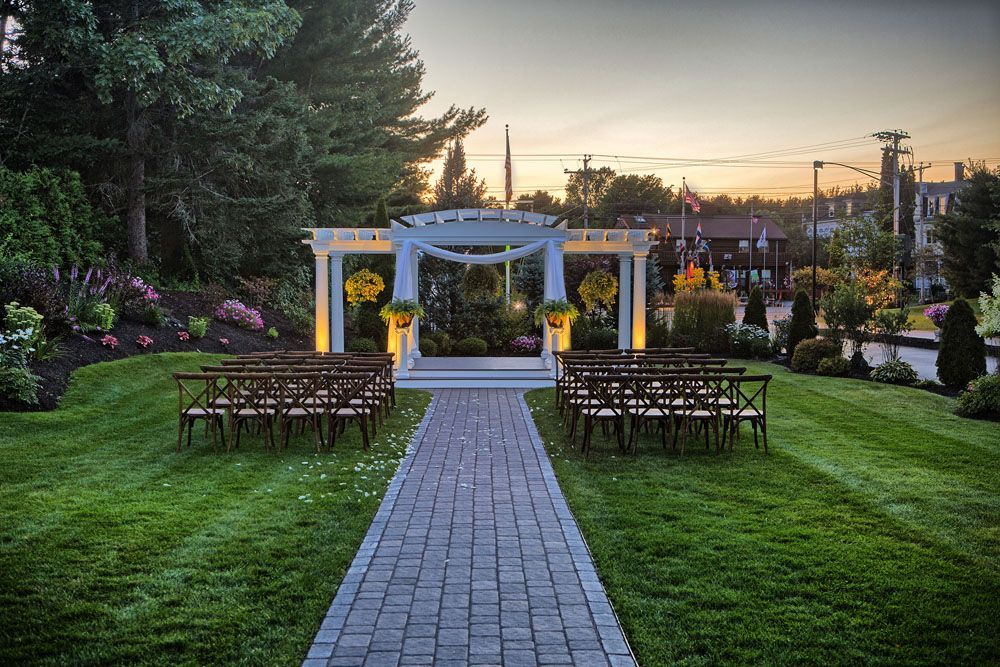 Wedding ceremony setup: white pergola, rows of chairs, brick path on green lawn, sunset backdrop.