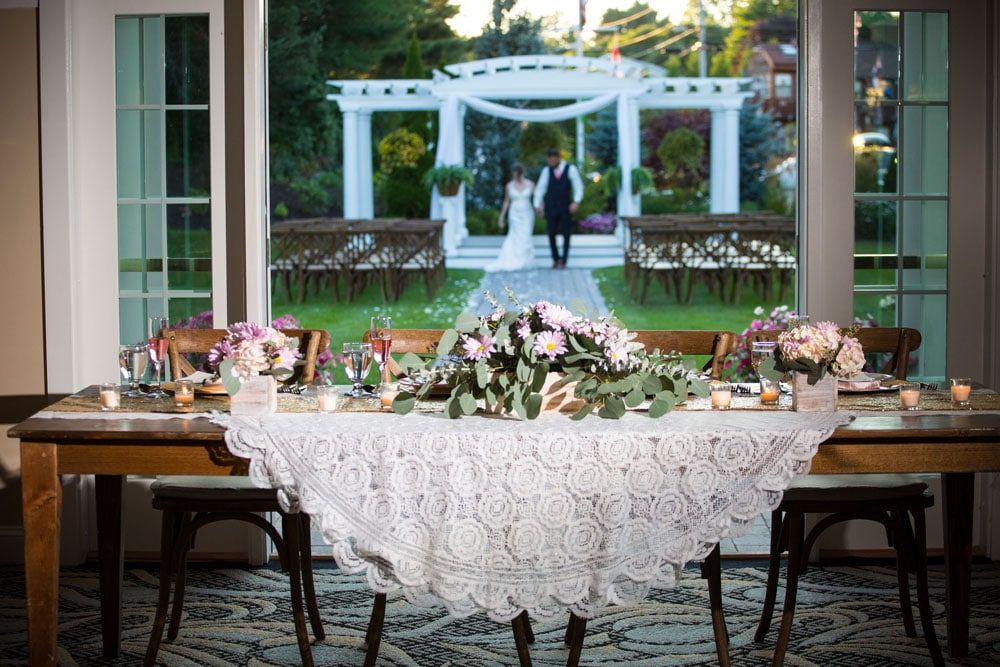 Wedding reception table with flowers, lace tablecloth, and blurred couple walking toward arbor outdoors.