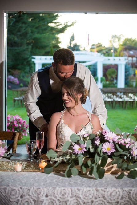 Groom kisses bride's head at wedding reception table with floral arrangement; outdoor setting with greenery.