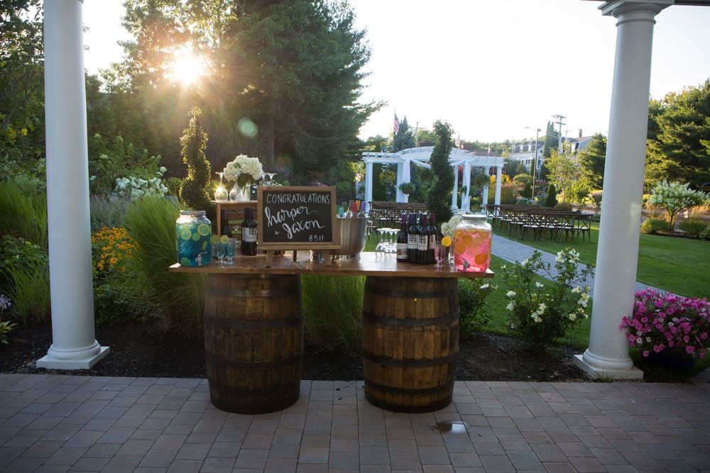 Outdoor bar with barrel base, drinks, and chalkboard sign, set against a garden and sunset.