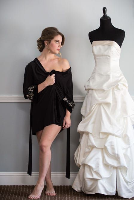 Bride in black robe looks at wedding dress on mannequin, standing in a room.