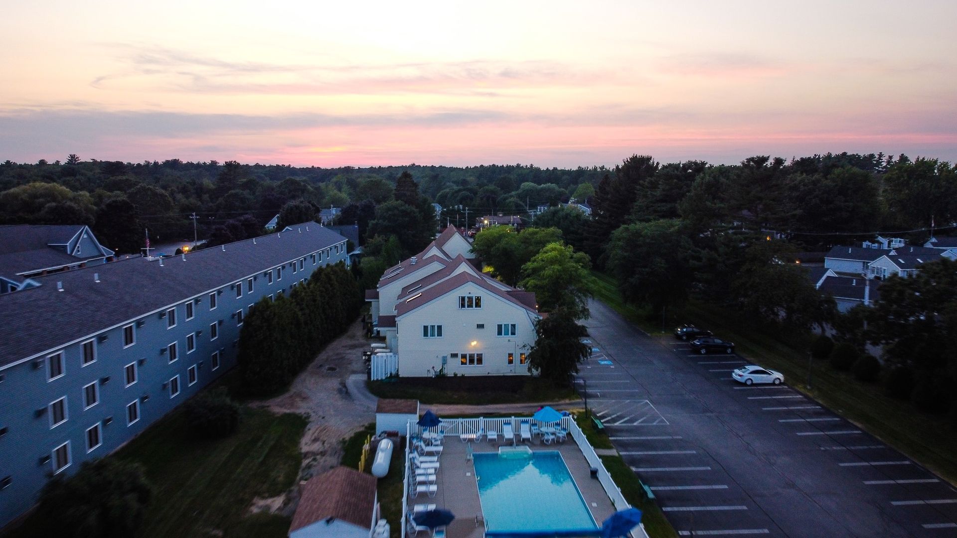 Aerial view of a neighborhood at dusk, with a pool, houses, parking lot, and trees.