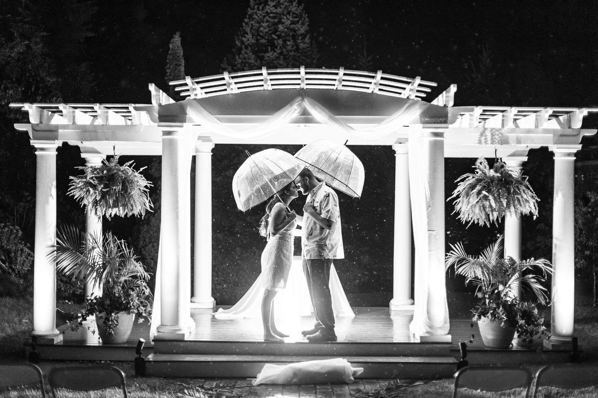 Bride and groom kissing under umbrellas during a rainy outdoor wedding ceremony beneath a decorated gazebo.