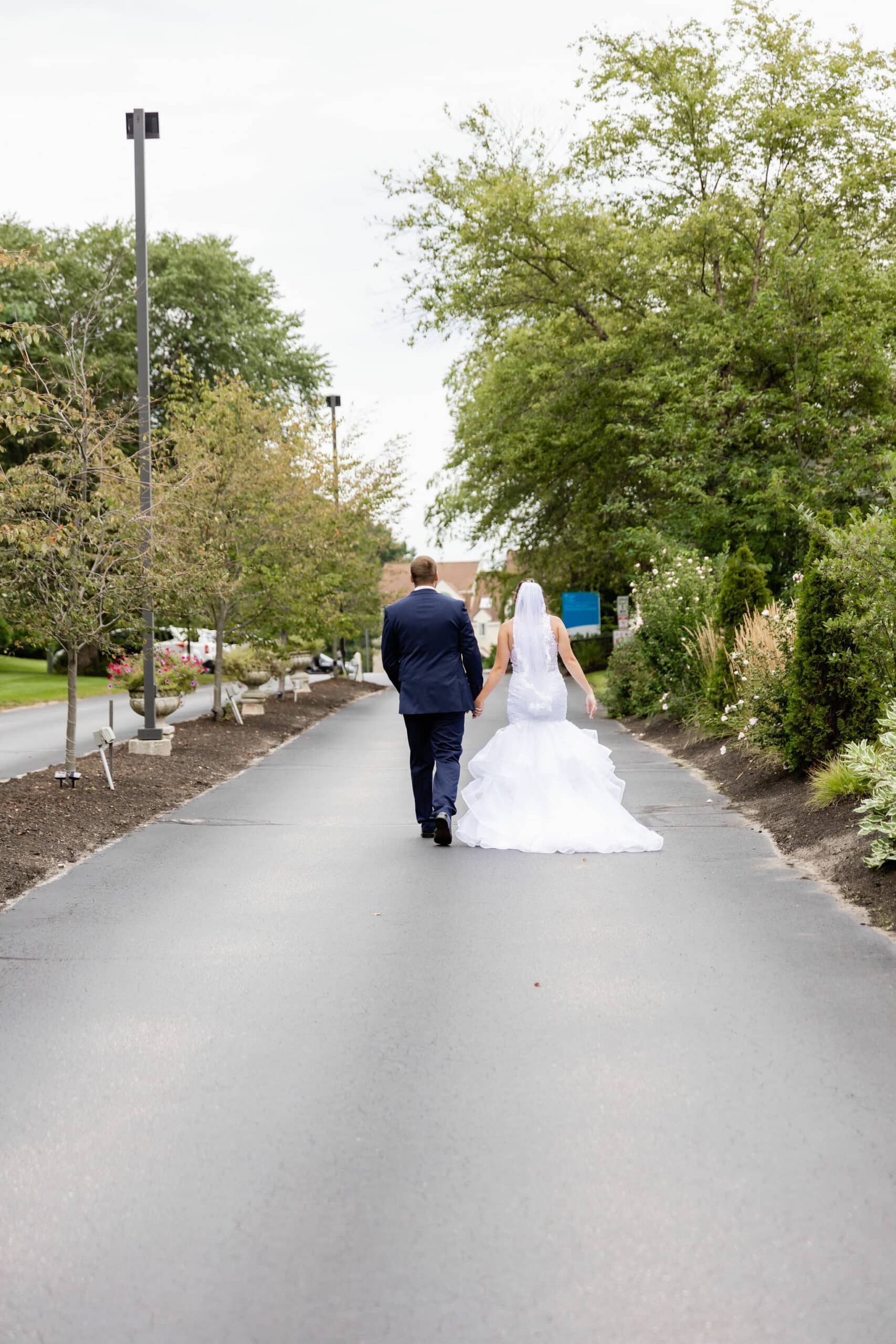 Bride and groom walk away on paved path, groom in navy suit, bride in white dress and veil, surrounded by trees.