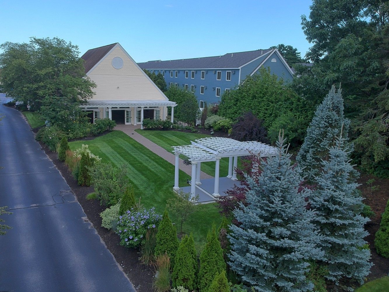 Roadside view of a building with a white pergola, trimmed lawn, and blue building behind it.