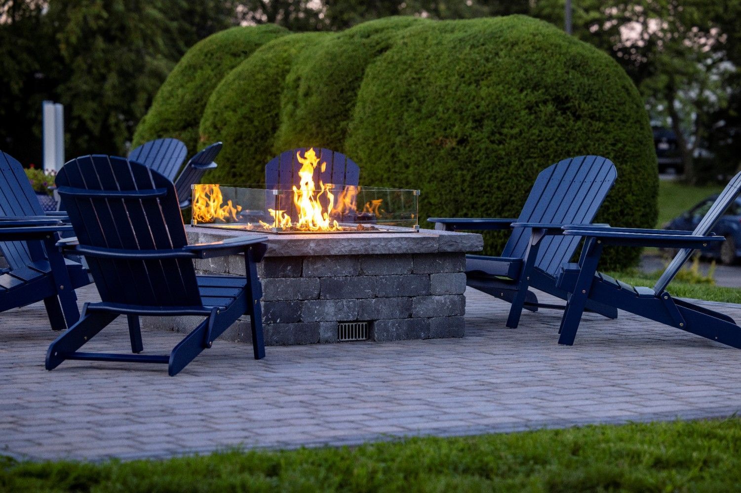 Blue chairs around a lit fire pit on a patio with a large green bush in the background.