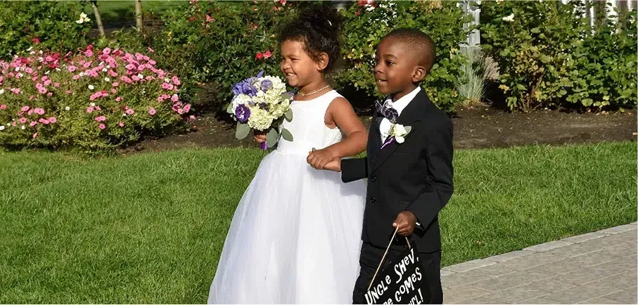 A flower girl and ring bearer holding hands, walking outside on a sunny day.