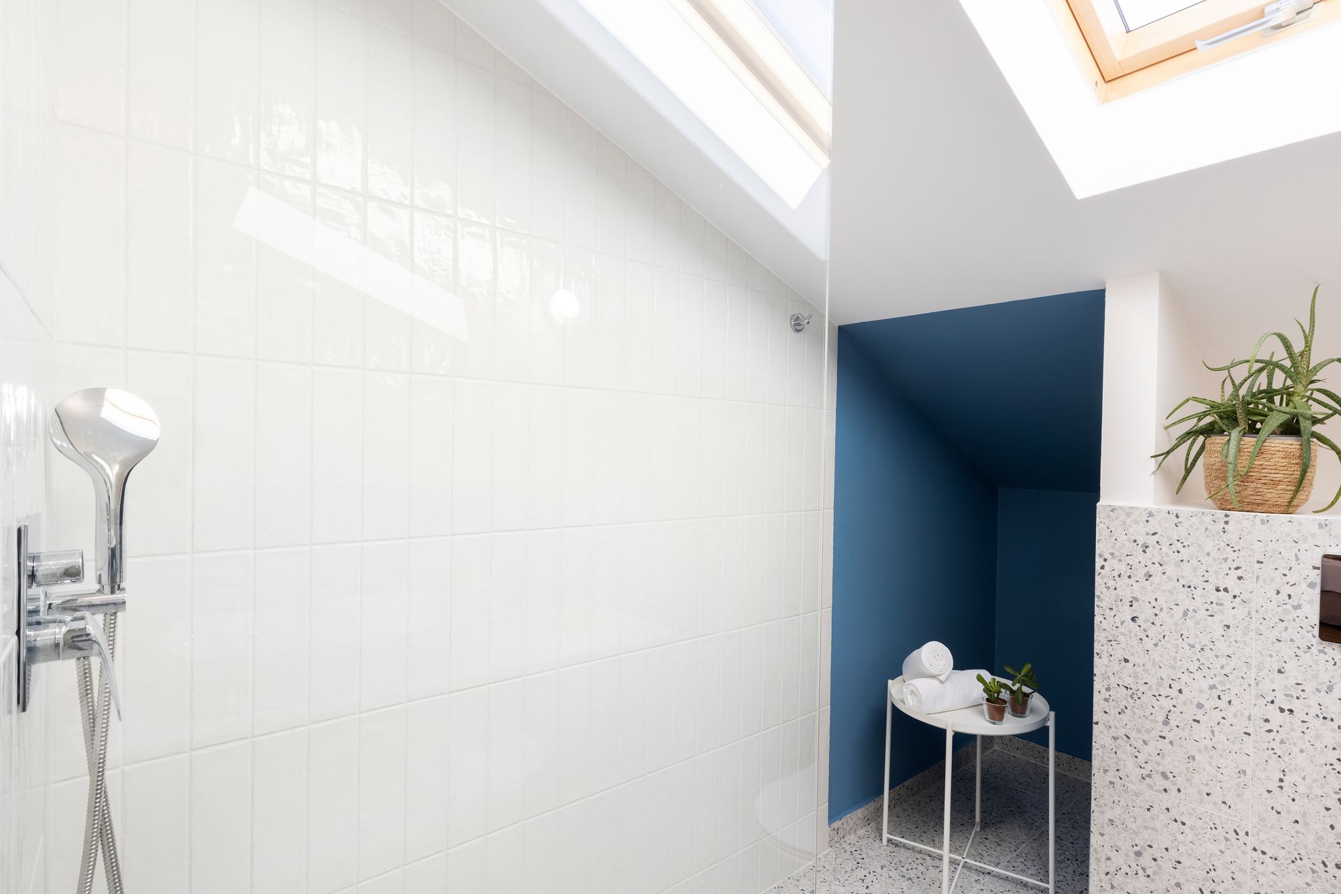 A bathroom with white tiles , a shower , a table and a skylight.
