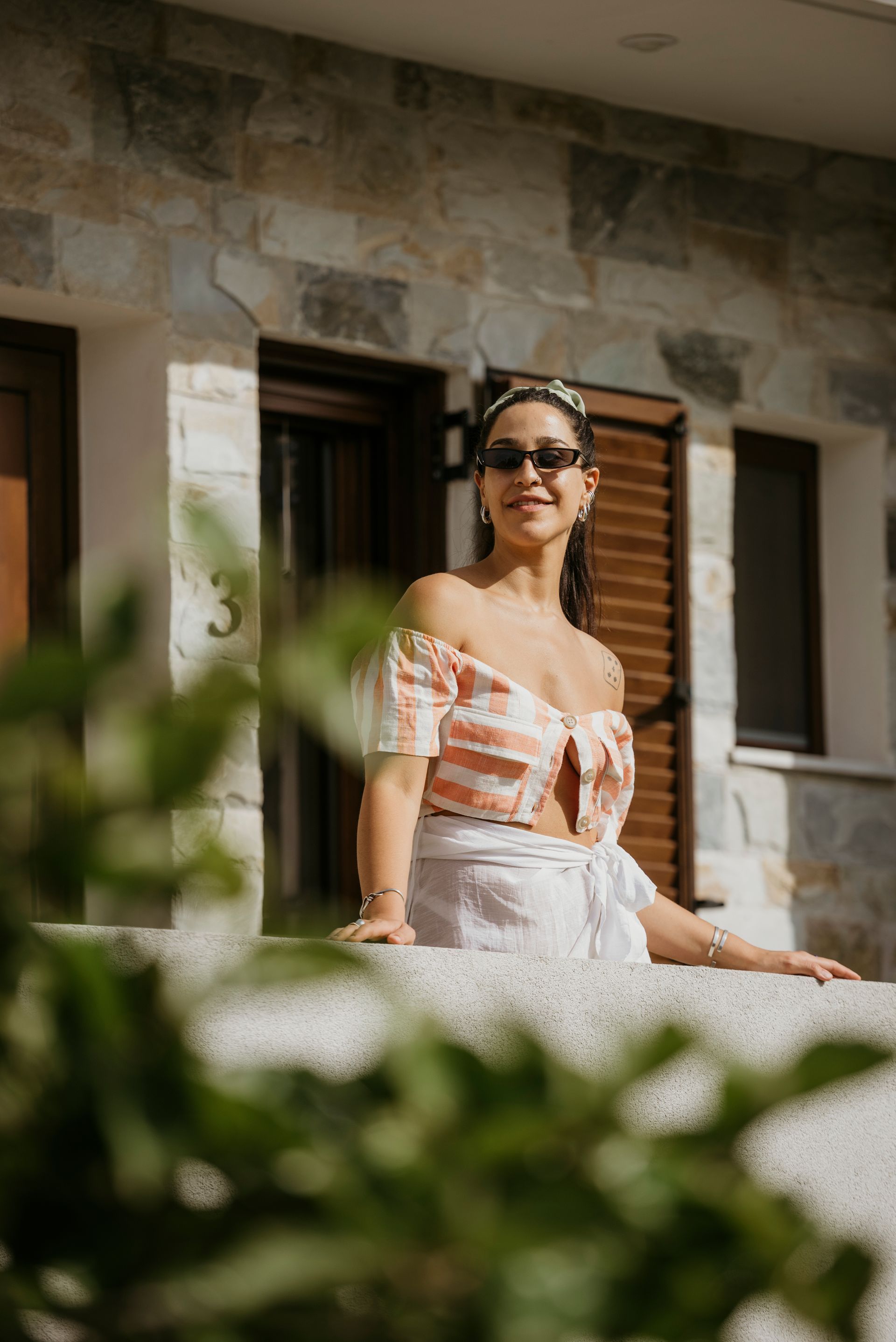 A woman wearing sunglasses is sitting on a balcony in front of a building.