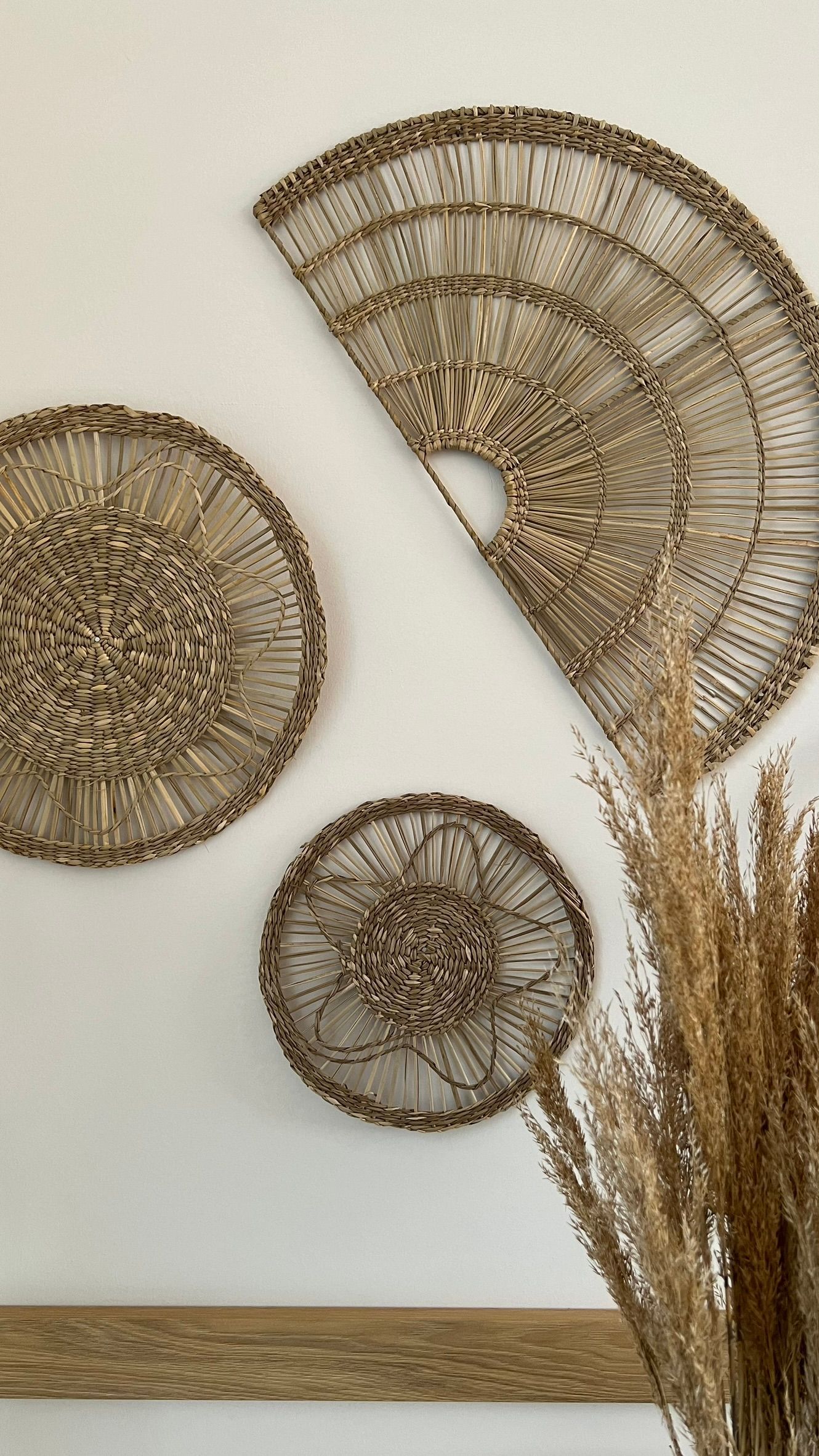 Three wicker fans are hanging on a wall next to a vase of dried grass.