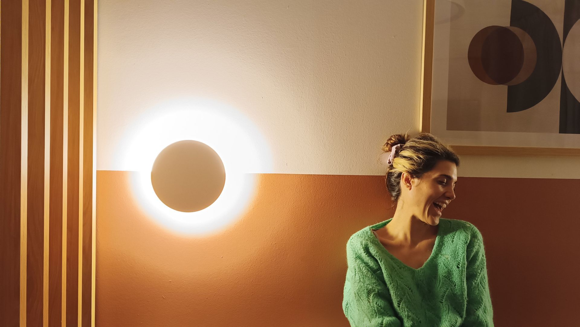 A woman in a green sweater is sitting in front of a wall with a light on it.