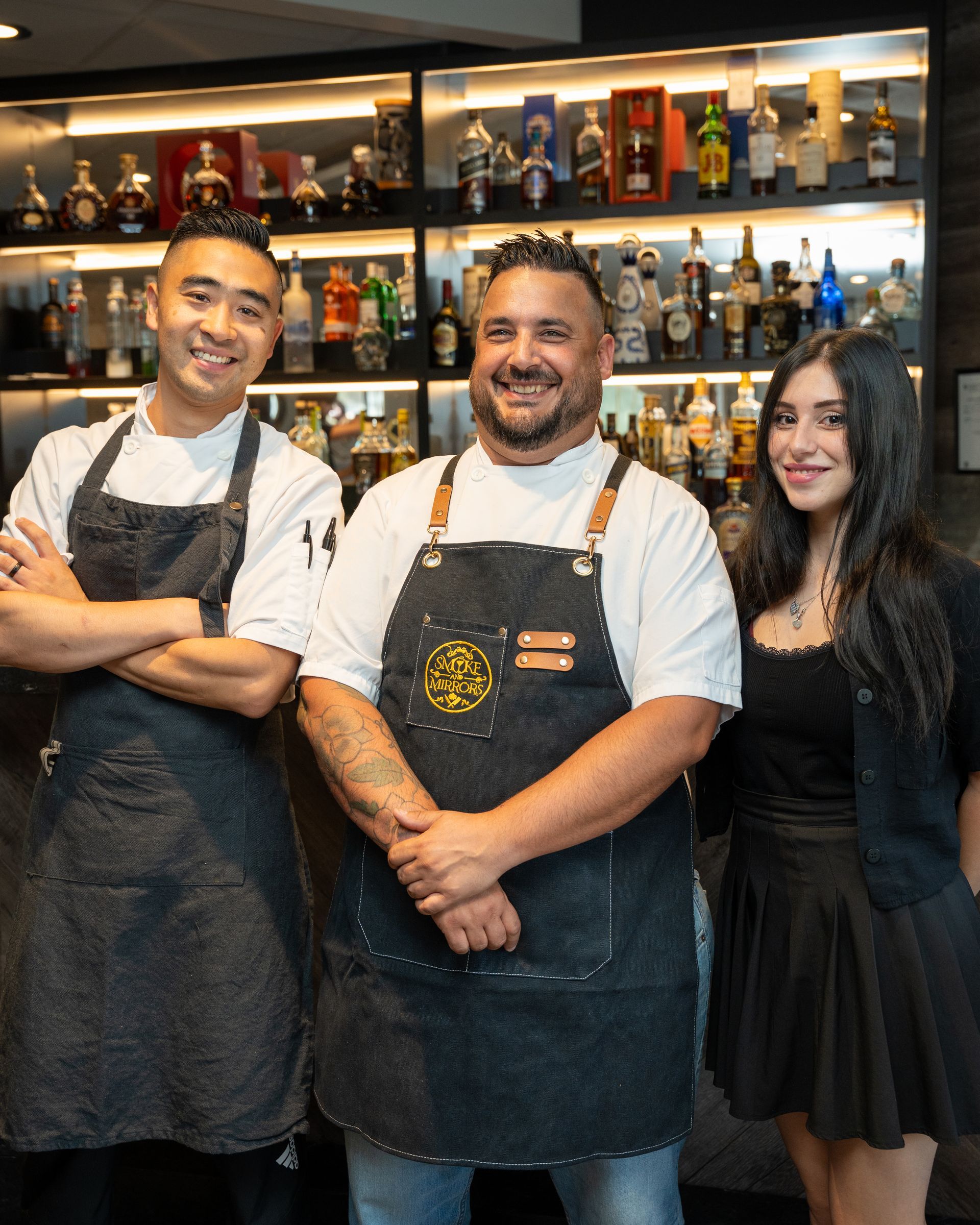Three restaurant employees smiling in front of a bar. One in center wearing black apron, others in white shirts and aprons.