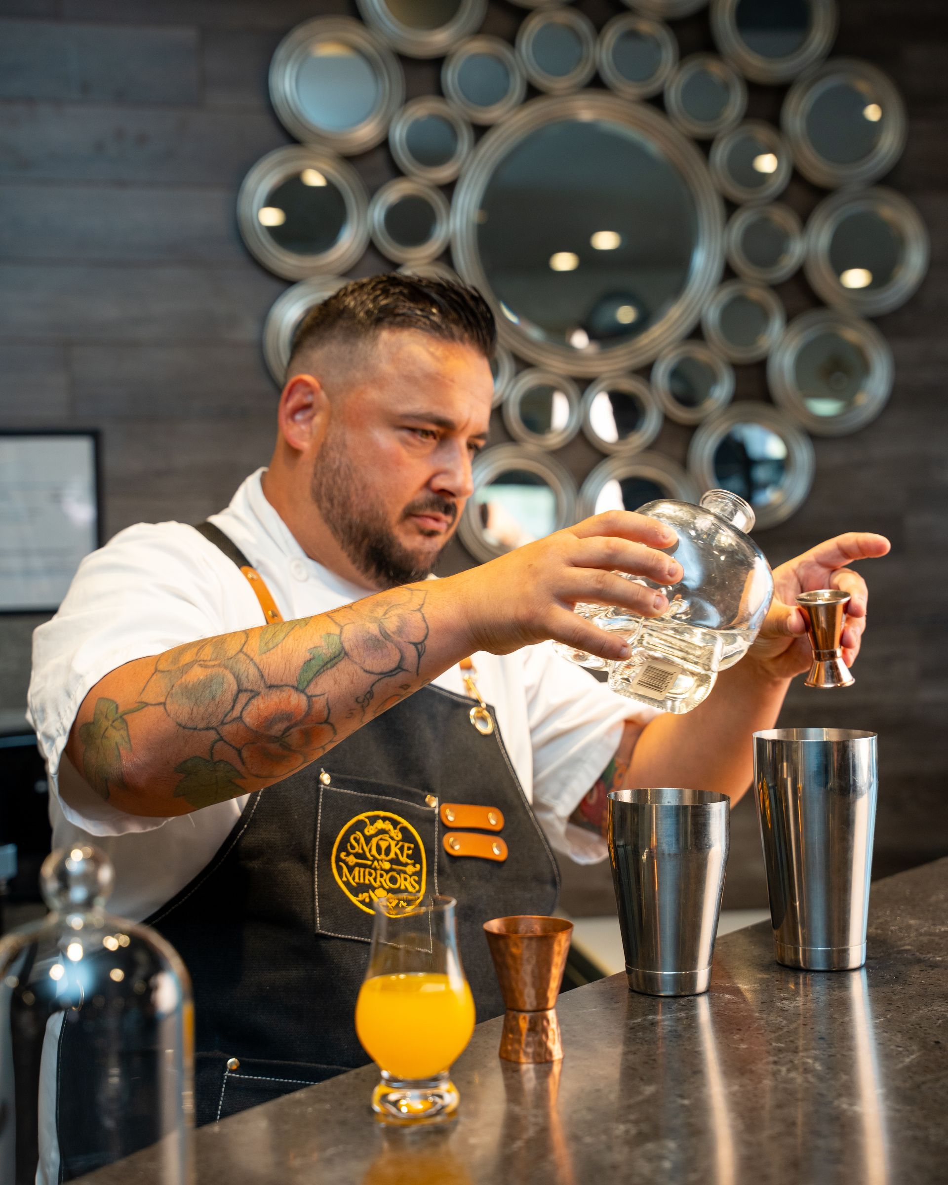 Bartender pouring cocktail into shaker; bar setting, tattoos, serious expression.