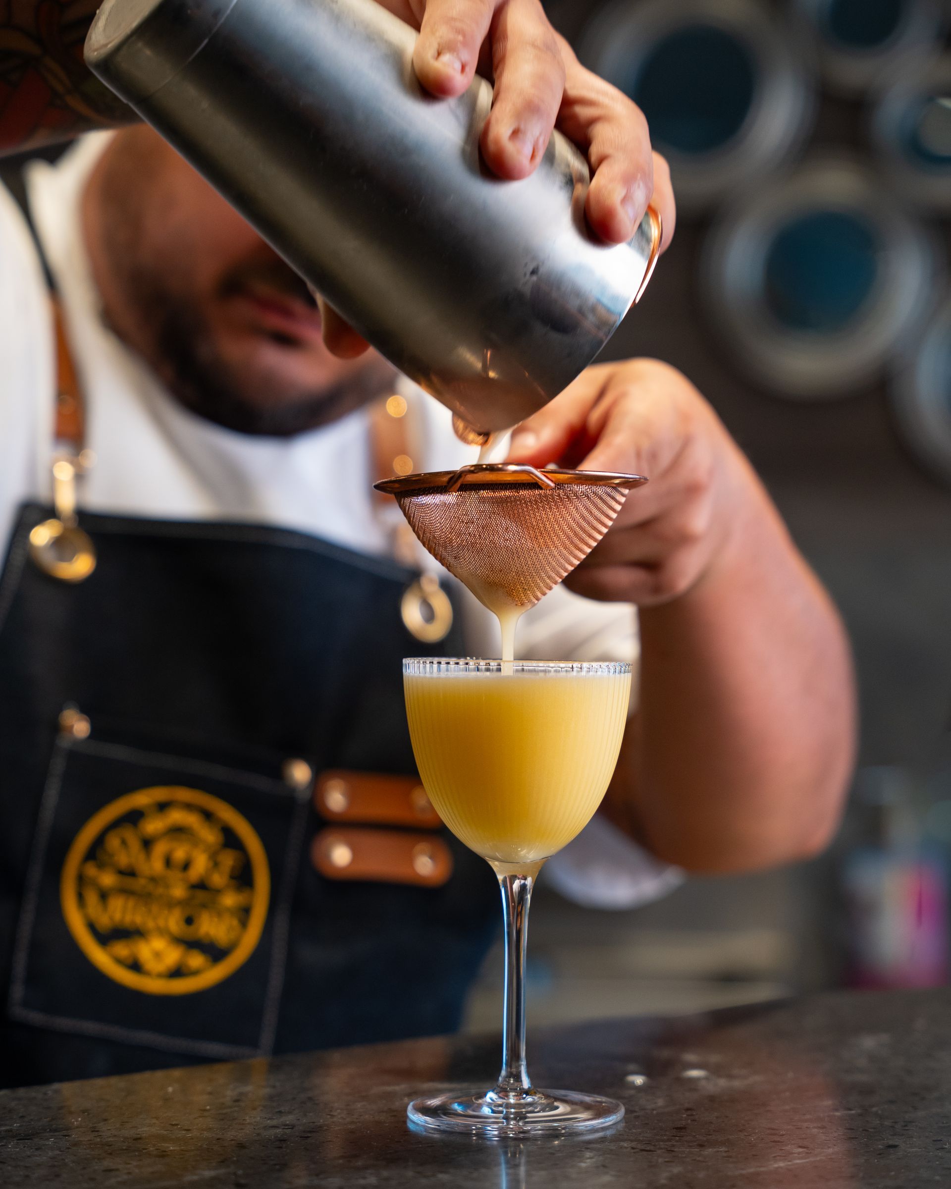 Bartender straining a yellow cocktail into a stemmed glass. Dark blue apron, brown leather straps, and metal shaker.
