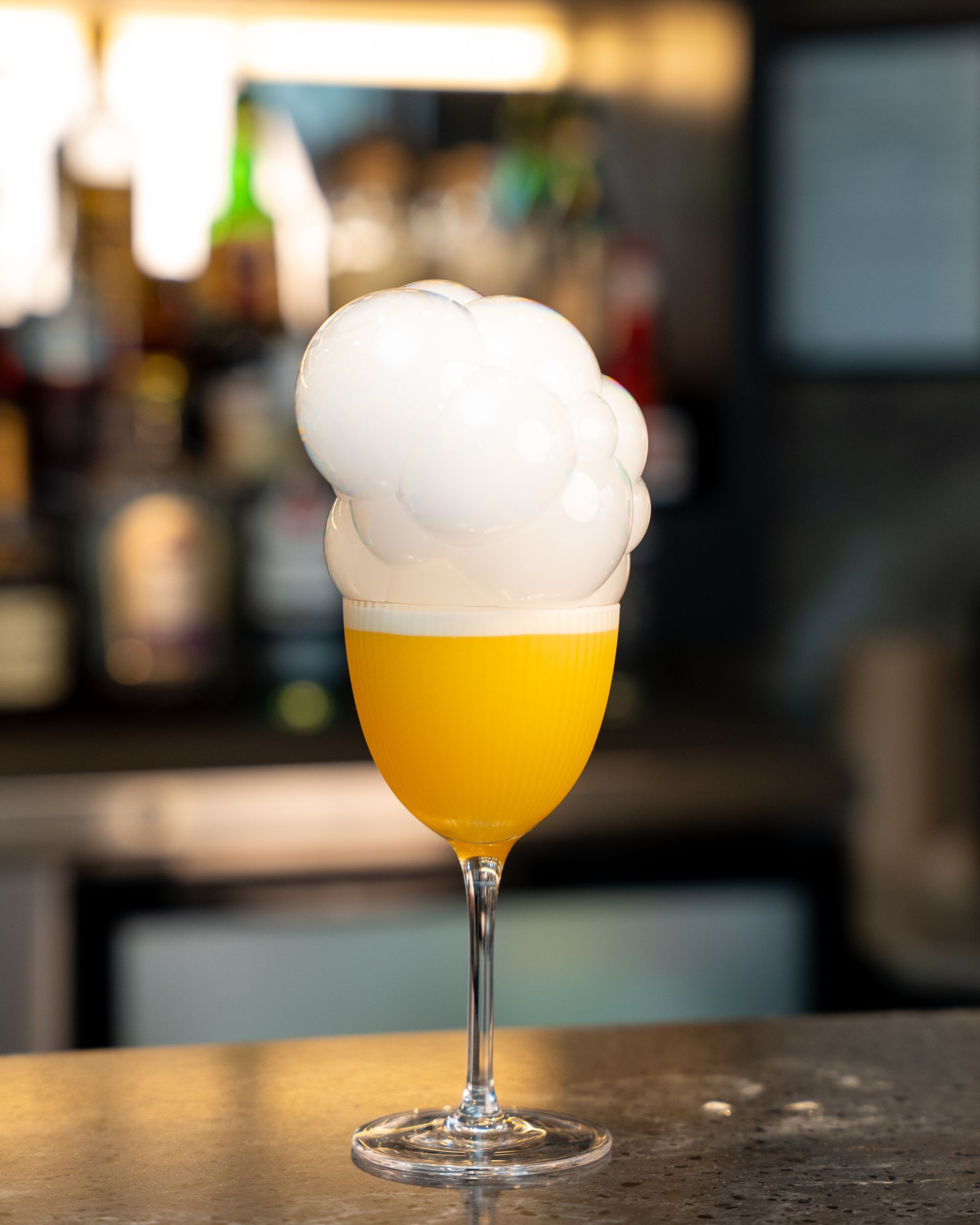 Yellow cocktail in a stemmed glass topped with white foam, on a bar.