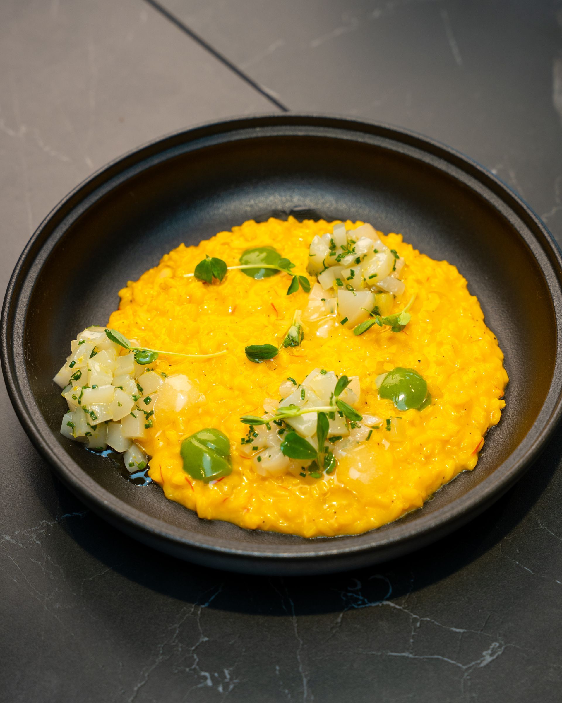 Orange risotto dish with white and green garnishes in a black bowl on a dark surface.