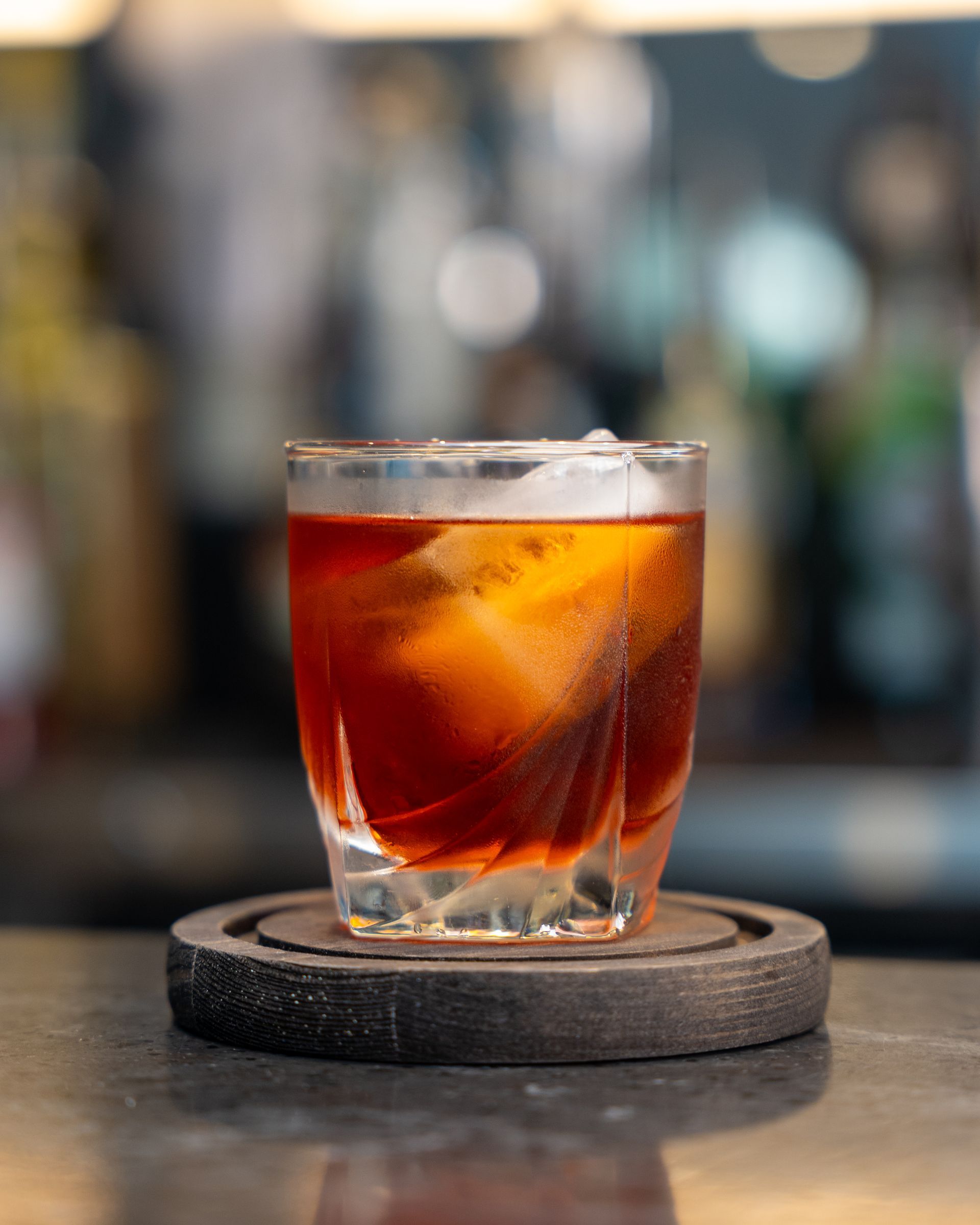 Dark cocktail in a rocks glass with ice, sitting on a coaster at a bar.