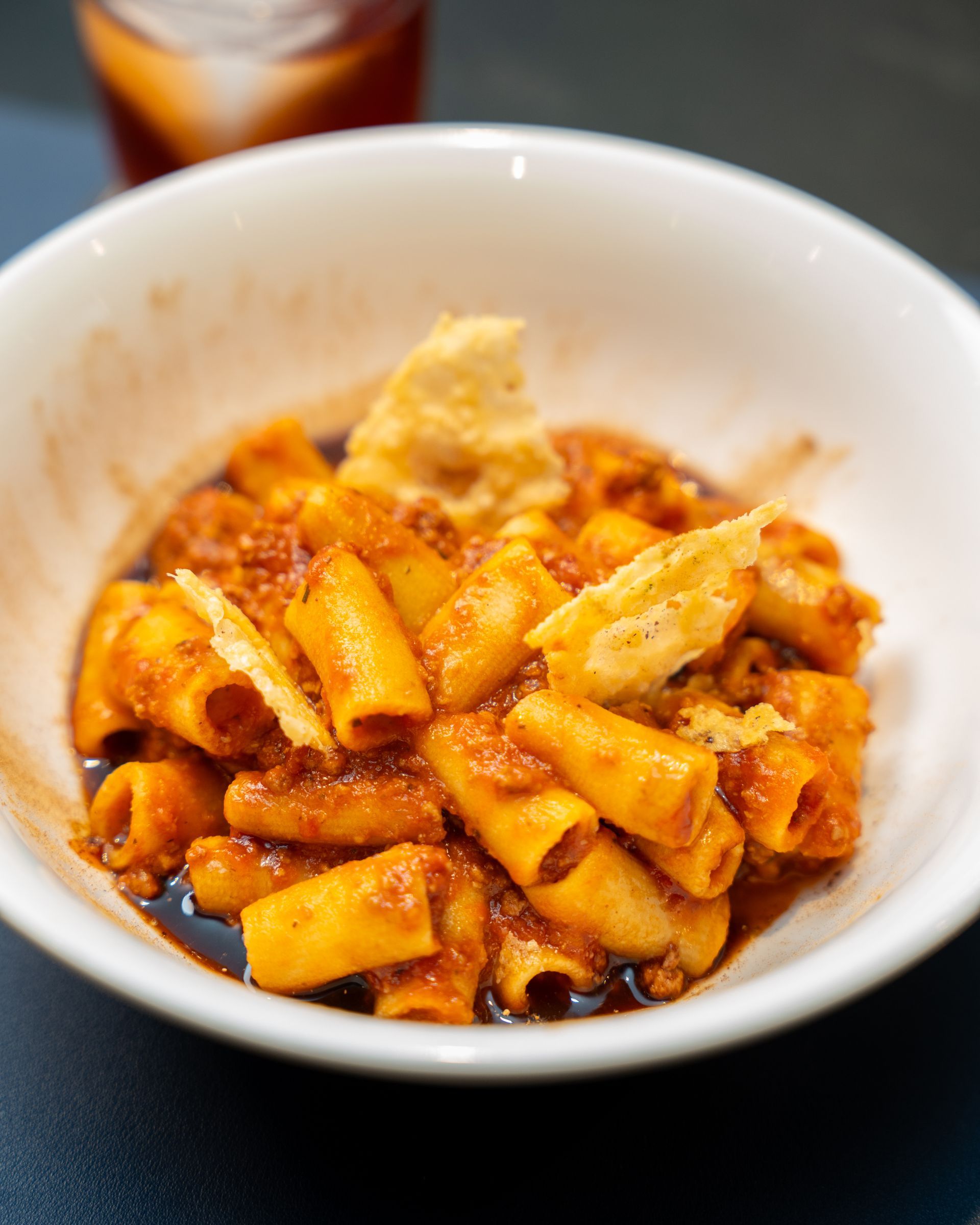 White bowl of rigatoni pasta with red sauce, topped with cheese, a dark drink in background.