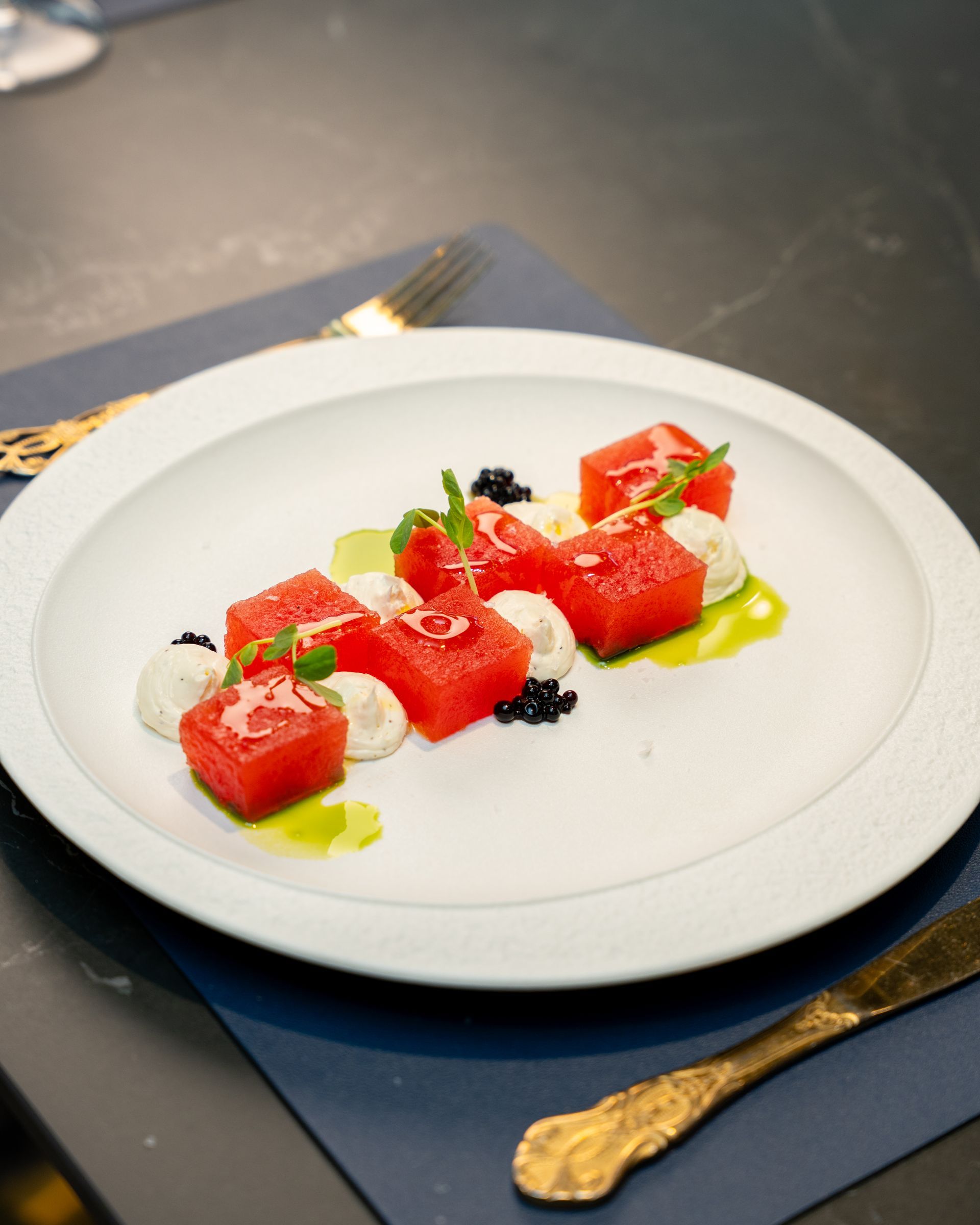 Watermelon cubes on a white plate with white cheese, green sauce, and dark caviar, with gold cutlery.