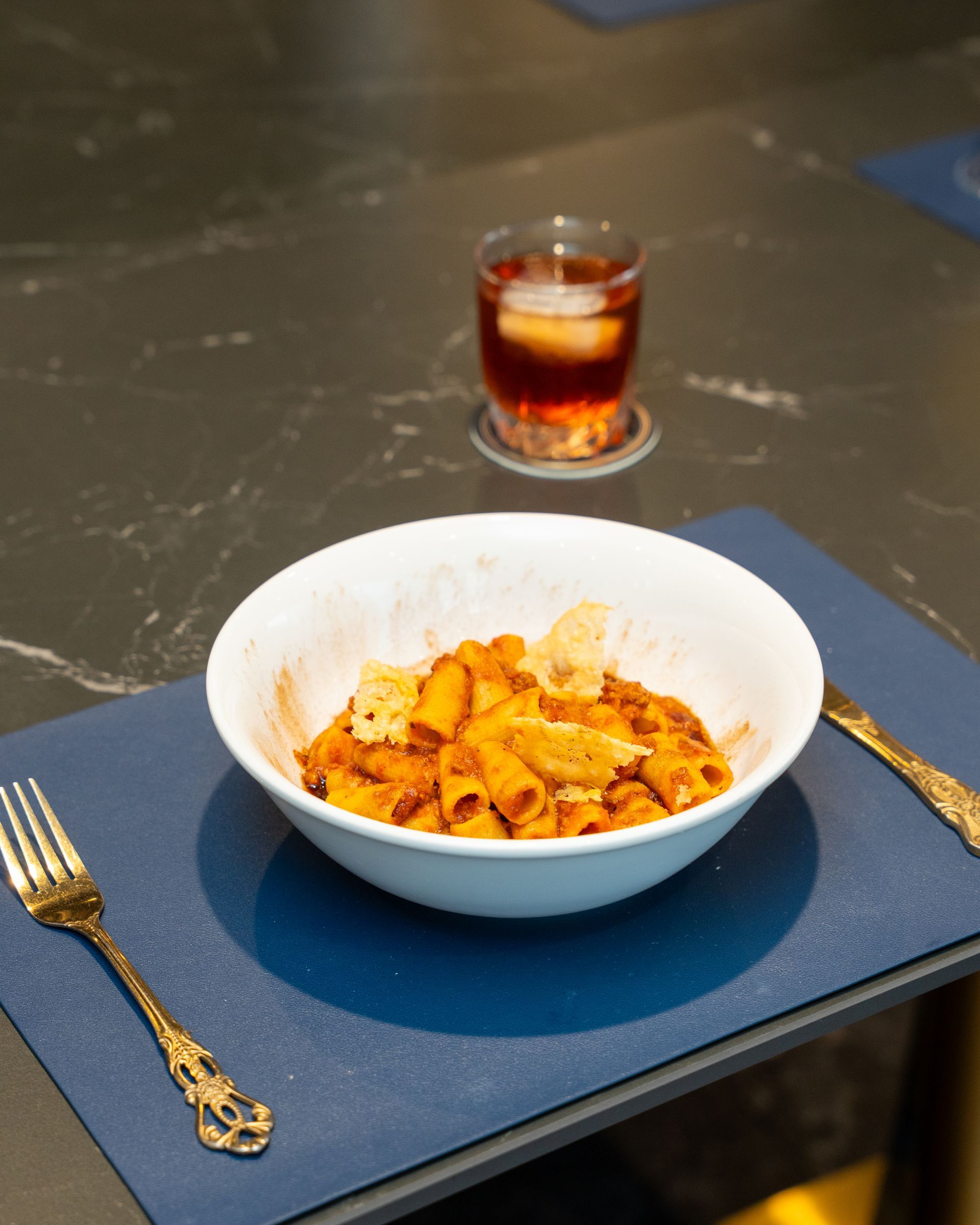 Pasta with red sauce, parmesan, and drink on a blue placemat with gold cutlery.
