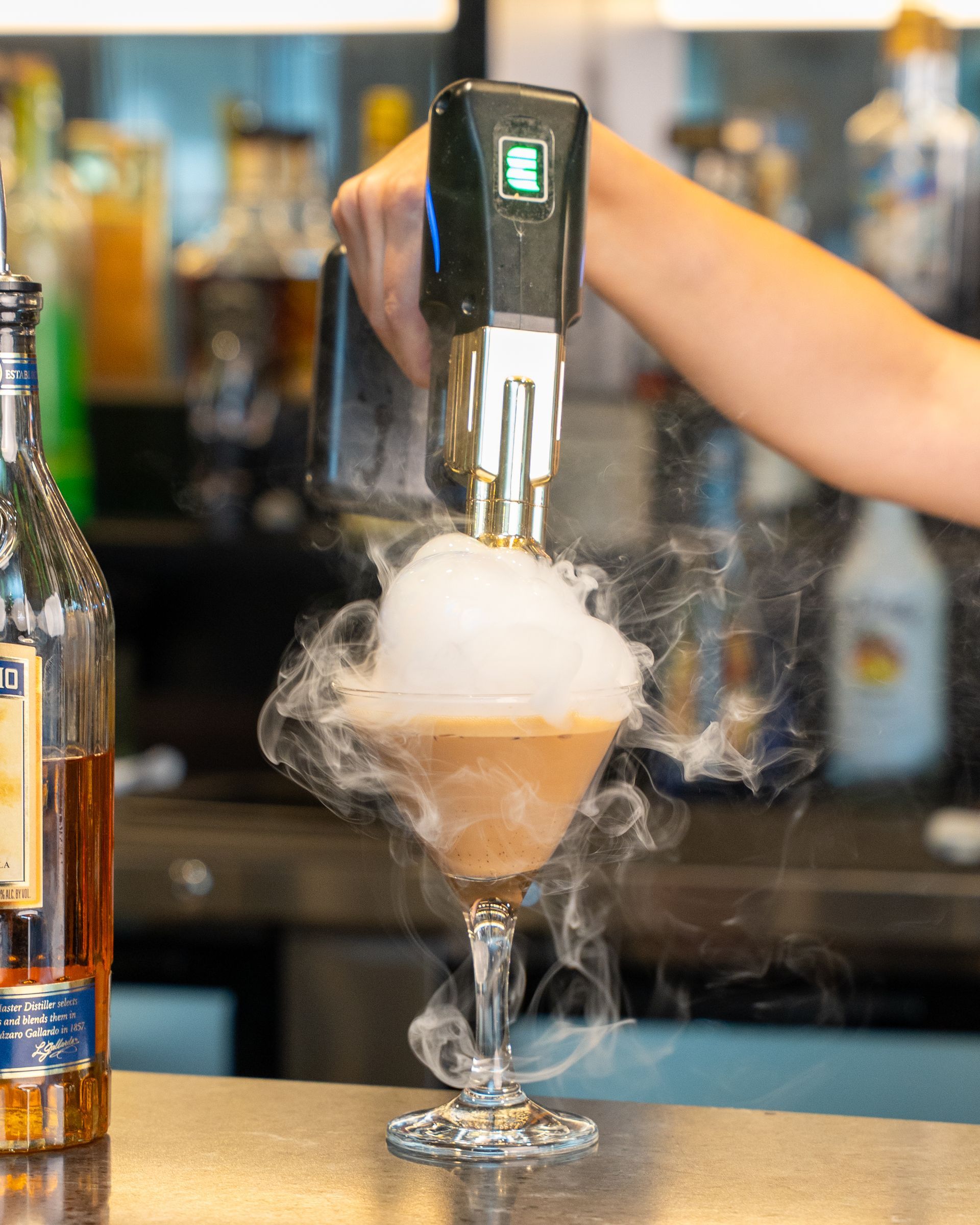 A bartender smokes a brown cocktail in a martini glass at a bar. Smoke billows around the glass.