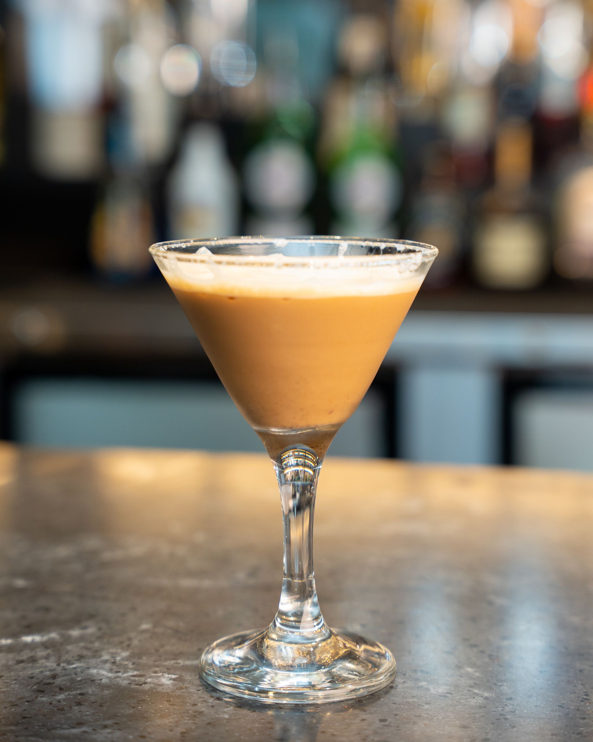 Cocktail in a martini glass on a bar. Light brown drink, sugar rim, blurred bar background.
