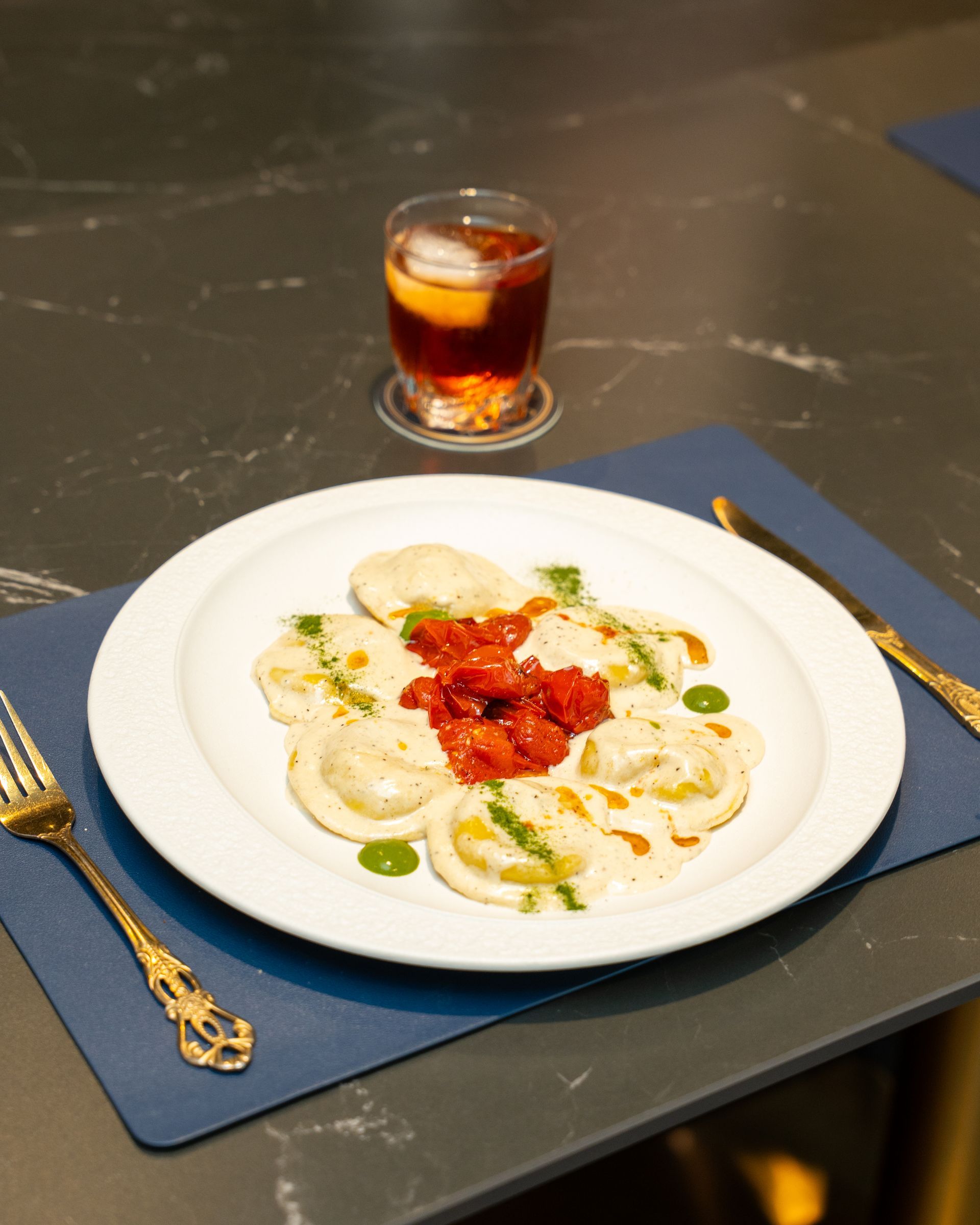 Plate of ravioli with a tomato topping, a cocktail, and silverware on a blue placemat.