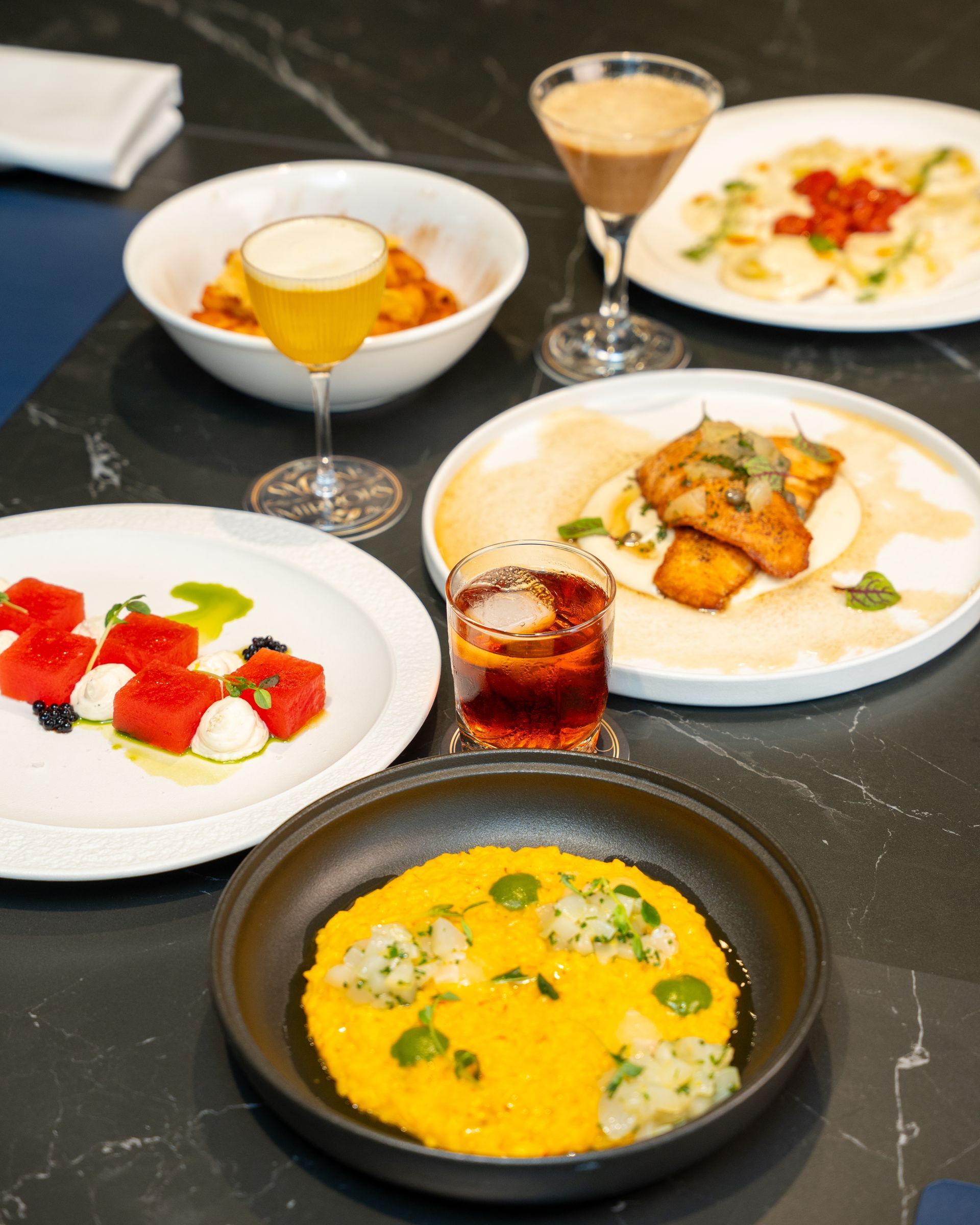 Multiple dishes and drinks on a dark table: watermelon salad, pasta, risotto, cocktails, fried food, and tacos.