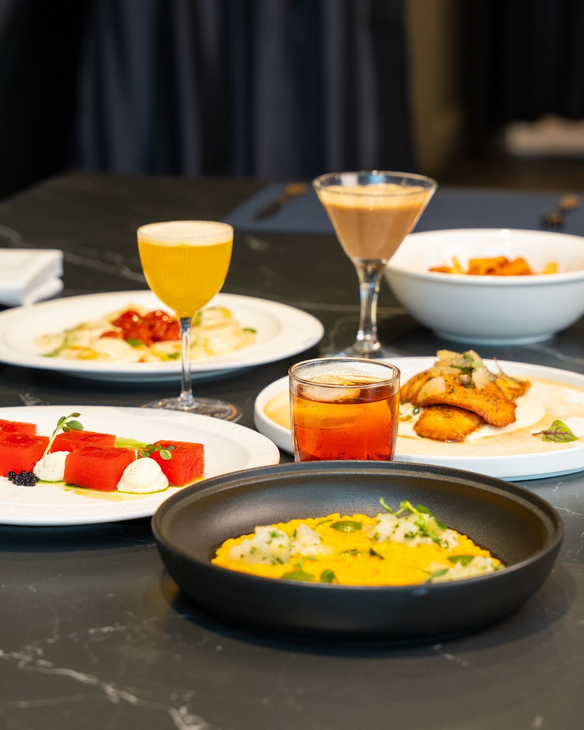 Table set for a meal: plates of food, drinks, and cocktails on a dark table.
