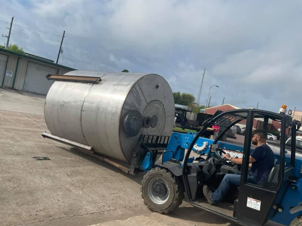 A man is driving a forklift carrying a large stainless steel tank.