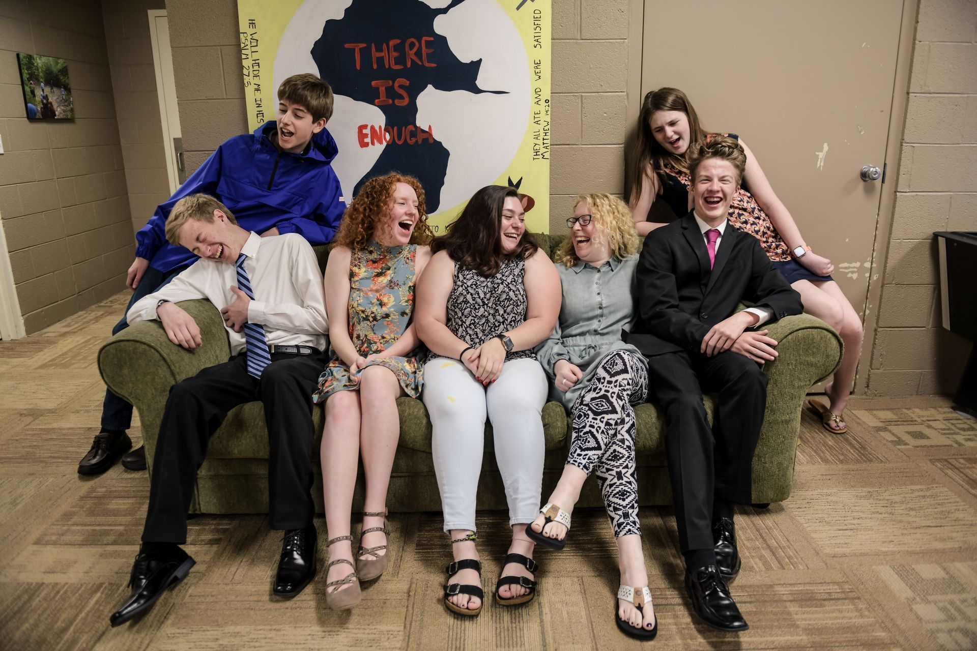 A group of seven people laughing on a green couch. A poster with