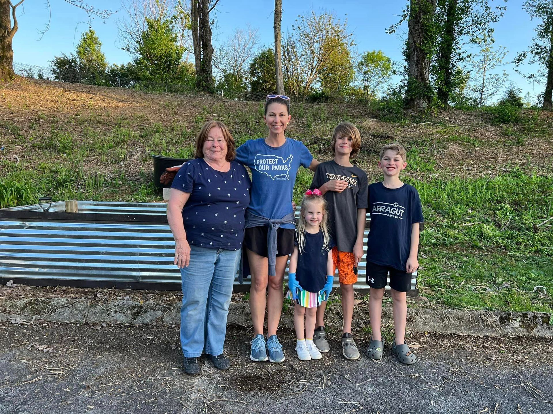 A group of five people smiling outdoors. A woman and four children stand in front of a weathered metal structure.