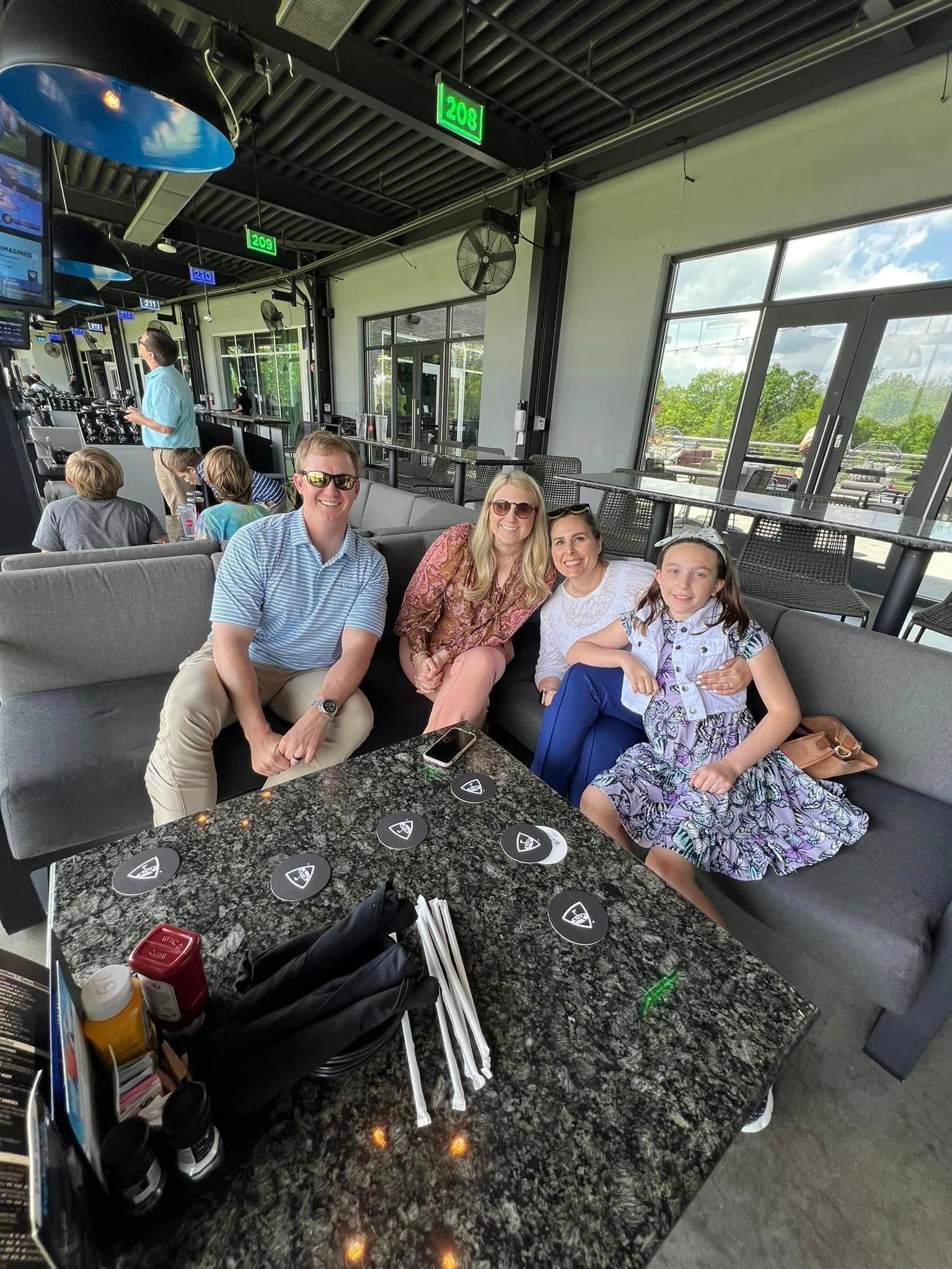 Group of four people at a Topgolf-like venue, sitting on a grey couch at a dark granite table.