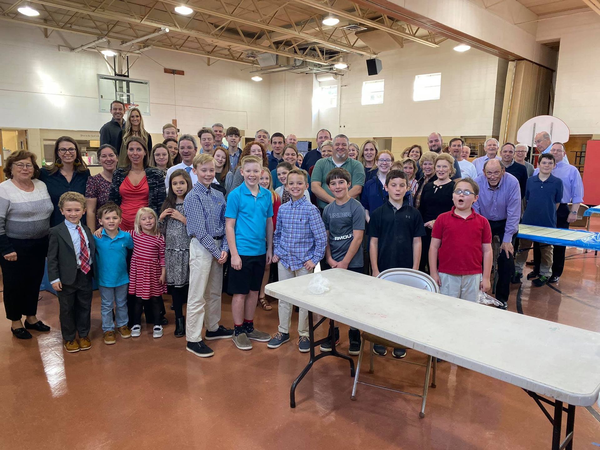 Large group of people, including children, gathered indoors for an event. They stand in a gymnasium with a long table in the foreground.