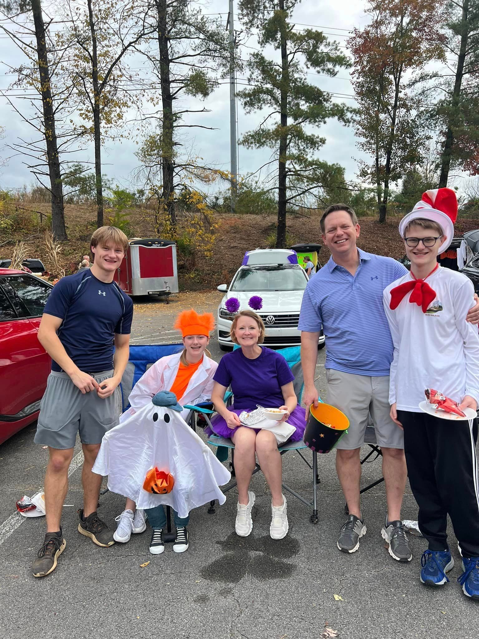 A group of five people dressed in costumes posing for a photo in front of a car decorated with purple balloons. Autumn setting.