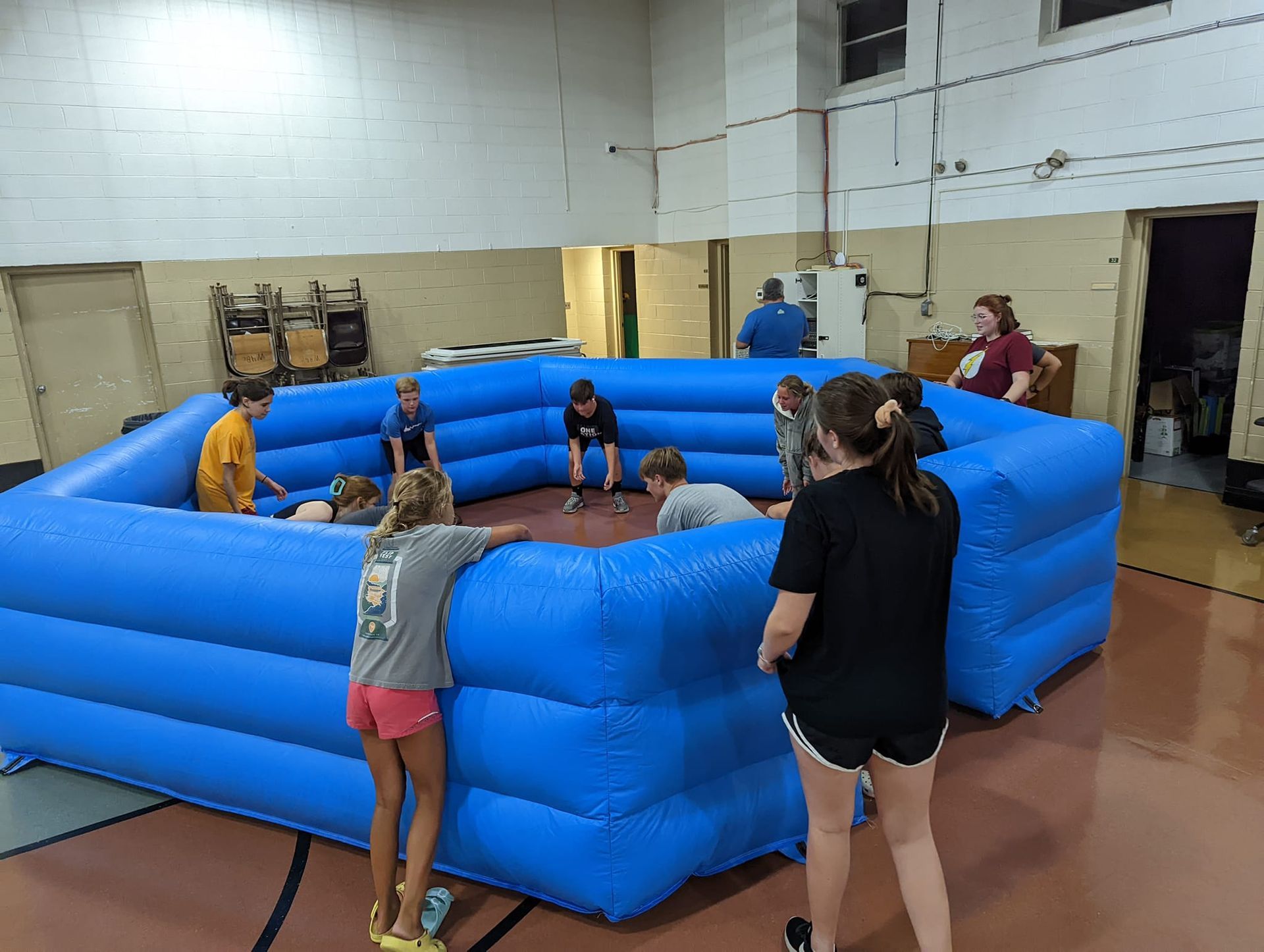 Children playing in a large, blue inflatable pit inside a gymnasium. Several kids stand around the perimeter, while others compete inside.