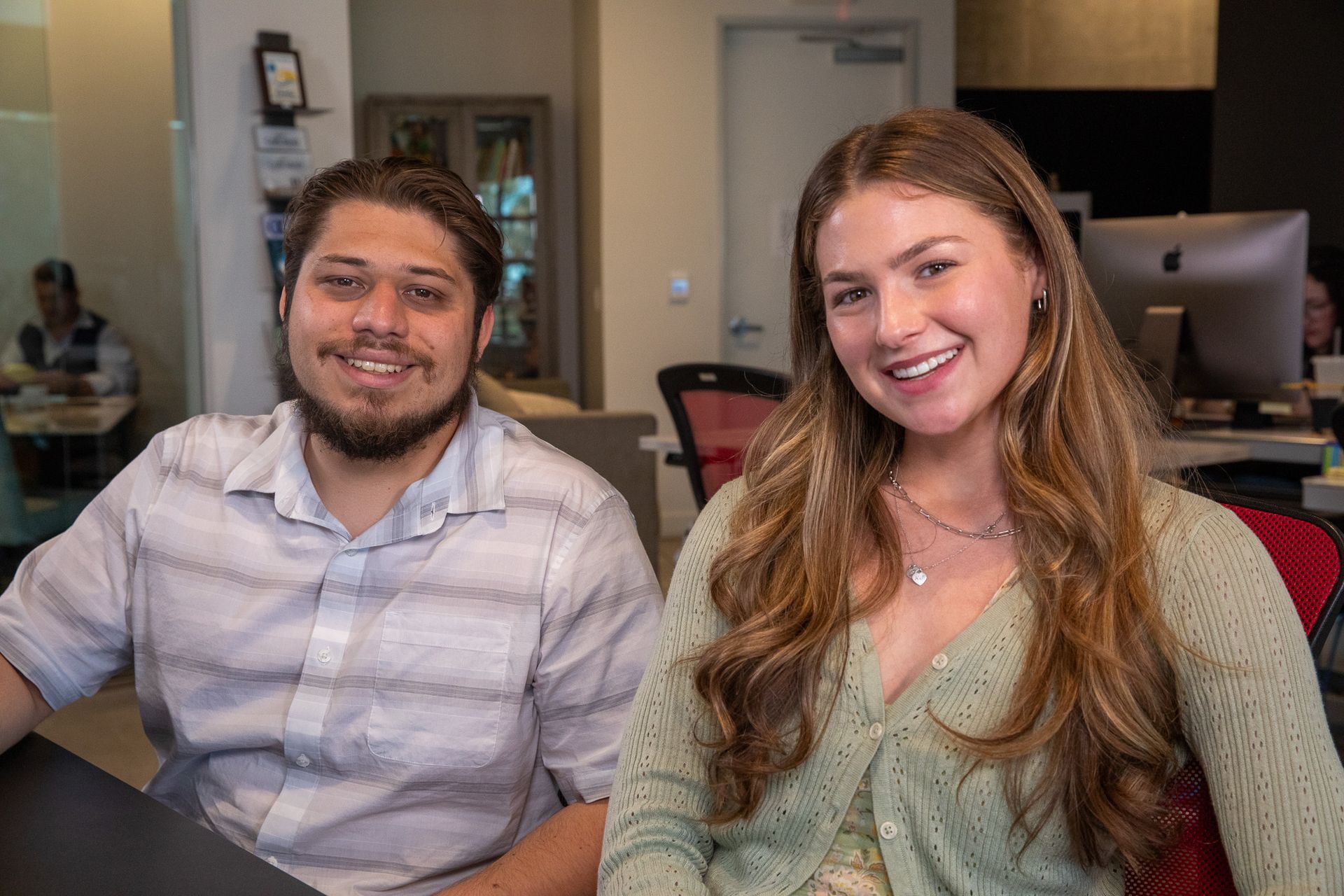 Two smiling young adults in an office setting; man with beard, woman with long hair, both looking at the camera.