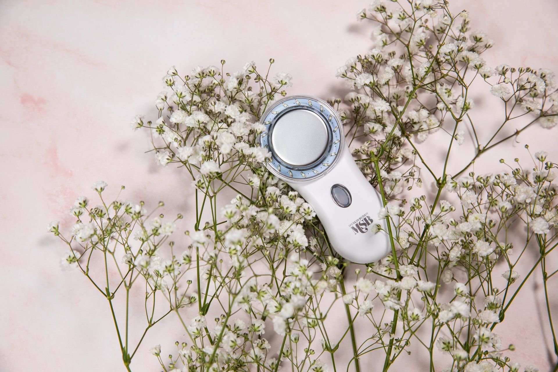 A white device is surrounded by white flowers on a pink surface.